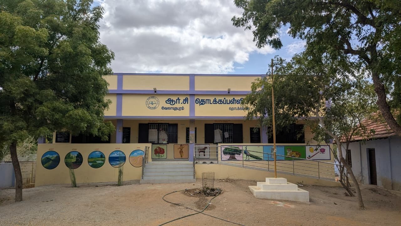 Completed single-storey primary school block at Velayudhapuram, painted cream with lilac pilasters, fronted by a muralled compound wall and a railed access ramp.